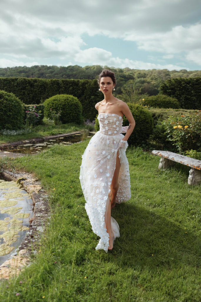 אישה בשמלת ערב לבנה שקופה ללא כתפיות עם חתך גבוה הולכת על הדשא בגינה עם גדר חיה מטופחת, ספסל אבן ובריכה תחת שמיים מעוננים. - Liel & Cavalli