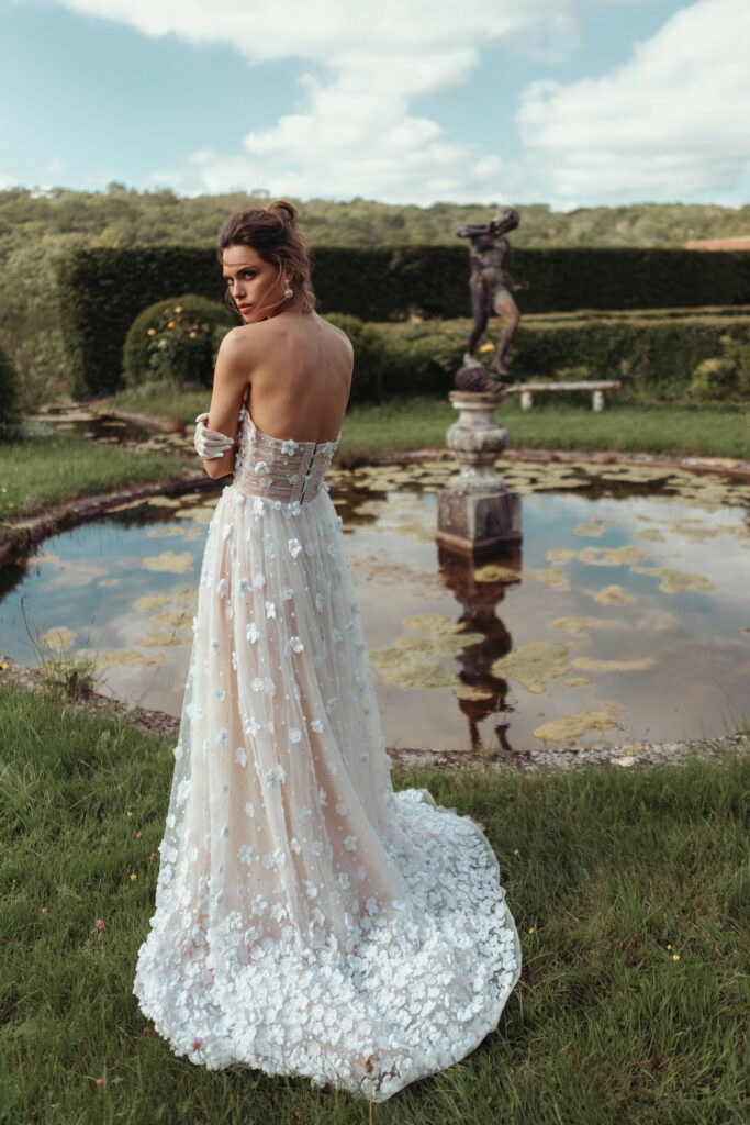 אישה בשמלה ללא כתפיות, רקומה בפרחים, עומדת מול בריכה עם פסל בגינה, מביטה מעבר לכתפה ביום מעונן חלקית. - Liel & Cavalli