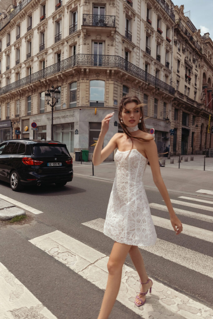 אישה לבושה בשמלת תחרה לבנה ללא כתפיות ונעלי עקב גבוהה הולכת ברחוב עירוני מול בניין אירופאי קלאסי עם מרפסות ופרטים מעוטרים. מכונית שחורה עוצרת במעבר החצייה מאחוריה. - Liel & Cavalli