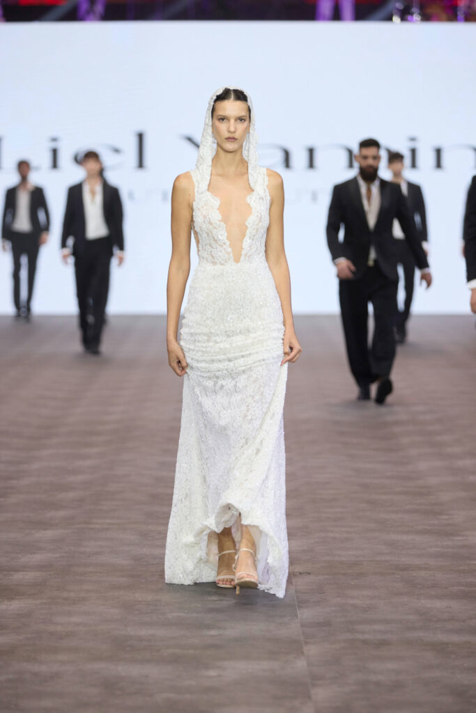דוגמנית צועדת על המסלול לבושה בשמלת תחרה לבנה ללא שרוולים, עם מחשוף V עמוק וכובע. כמה גברים בחליפות צועדים מאחוריה על מסלול תצוגת האופנה. - Liel & Cavalli