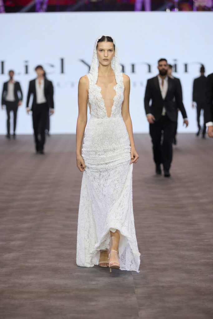 דוגמנית צועדת על המסלול בשמלת כלה לבנה, ללא שרוולים, עם מחשוף עמוק וצעיף תחרה תואם. ברקע נראים כמה אנשים לבושים בחליפות שחורות רשמיות. - Liel & Cavalli