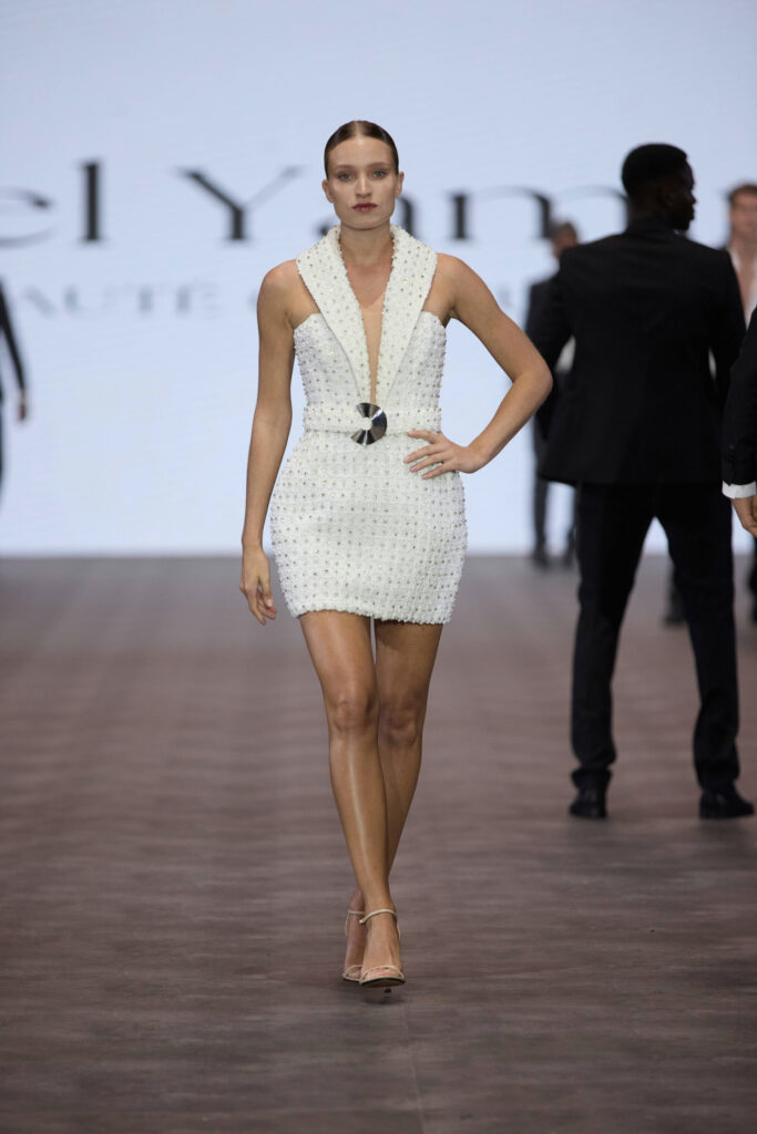 דוגמנית צועדת על מסלול תצוגה לבושה בשמלת מיני לבנה ללא שרוולים, עם מחשוף עמוק ואבזם עגול במותניים. יש לה שיער חלק ועקבים בצבע גוף. ברקע נראים אנשים אחרים צועדים. - Liel & Cavalli