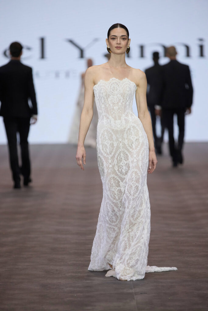 דוגמנית צועדת על המסלול בשמלת תחרה לבנה, צמודה לגוף, ללא כתפיות, עם שובל עדין. ברקע נראות דמויות מטושטשות ומסך גדול עם טקסט חלקי. - Liel & Cavalli