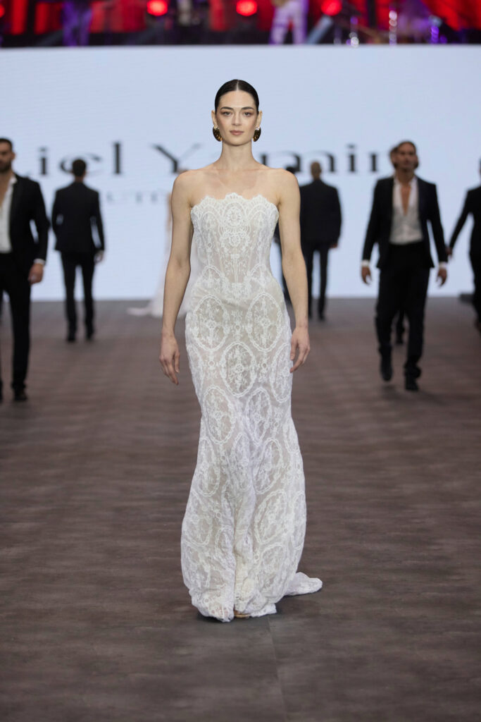 דוגמנית צועדת על מסלול תצוגה לבושה בשמלת תחרה לבנה צמודה ללא כתפיות, כשברקע מספר אנשים לבושים בחליפות שחורות ומאחוריה מסך מואר. - Liel & Cavalli