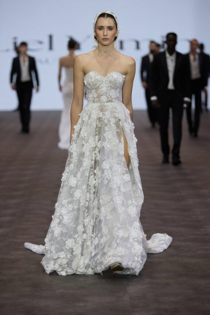 דוגמנית צועדת על מסלול תצוגה לבושה בשמלת כלה לבנה ללא כתפיות, מעוטרת בתחרה פרחונית, עם שובל ארוך. ברקע צועדים ארבעה דוגמנים נוספים לבושים בחליפות כהות וחולצות בהירות. - Liel & Cavalli