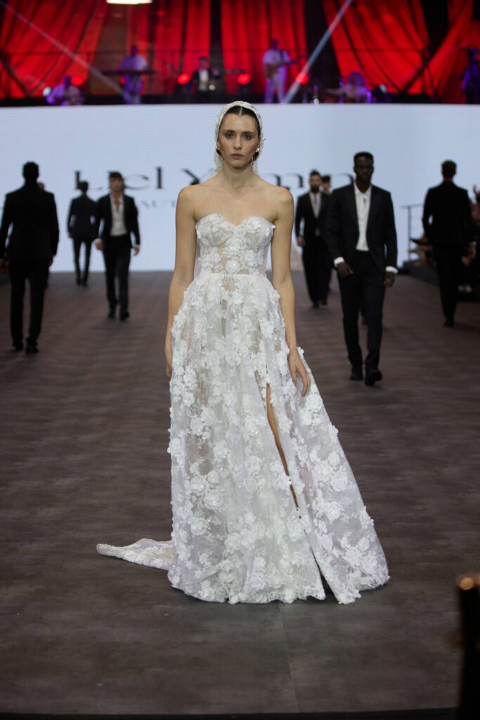דוגמנית צועדת על מסלול תצוגה לבושה בשמלת כלה לבנה ללא כתפיות, עם חתך גבוה עד הירך ודוגמאות פרחוניות. ברקע נראים כמה אנשים בחליפות כהות, ווילונות אדומים תלויים מעל הבמה. - Liel & Cavalli