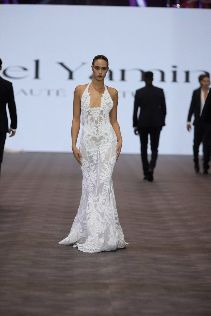 דוגמנית צועדת על המסלול לבושה בשמלה לבנה ללא שרוולים, עם דוגמת תחרה וצווארון הלטר. ברקע נראים כמה אנשים בחליפות כהות, ומסך גדול מציג טקסט מטושטש. - Liel & Cavalli