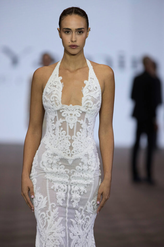 דוגמנית צועדת על מסלול תצוגה לבושה בשמלת סטרפלס לבנה ושקופה עם תחרה ועיטורים פרחוניים מורכבים; הרקע מטושטש ורואים בו צלליות של אנשים. - Liel & Cavalli