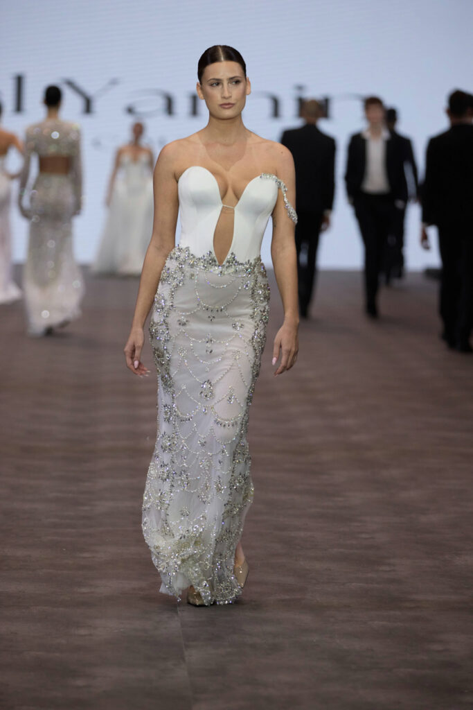 דוגמנית צועדת על המסלול בשמלה מעוטרת בלבן וכסף עם מחשוף עמוק וחצאית שקופה ומחוטבת בתצוגת אופנה, עם דמויות מטושטשות ורקע מואר ברקע. - Liel & Cavalli