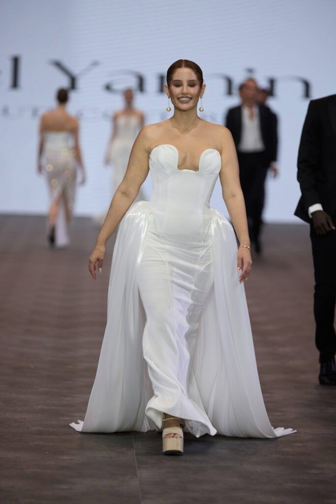 דוגמנית מחייכת לבושה בשמלת ערב לבנה ללא כתפיות עם מחוך צמוד ובד דרפטי צועדת בביטחון על המסלול, כשברקע דוגמניות אחרות ומסך ענק. - Liel & Cavalli