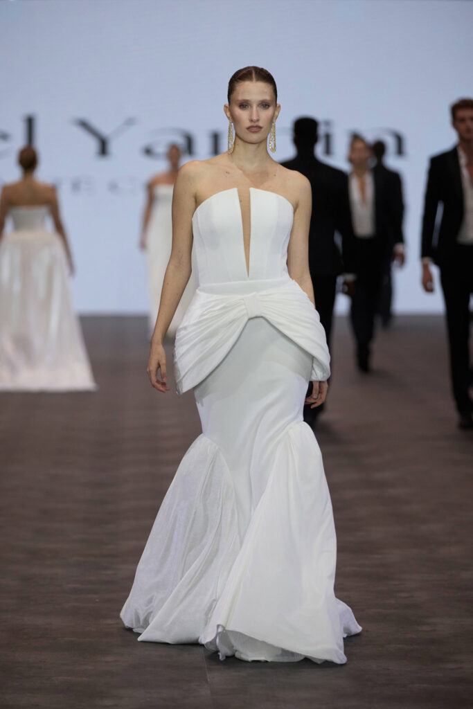 דוגמנית צועדת על המסלול לבושה בשמלת ערב לבנה ללא כתפיות עם פפלום מבנה וחצאית בסגנון בת ים. ברקע נראות דמויות מטושטשות ומסך עם טקסט חלקי. - Liel & Cavalli