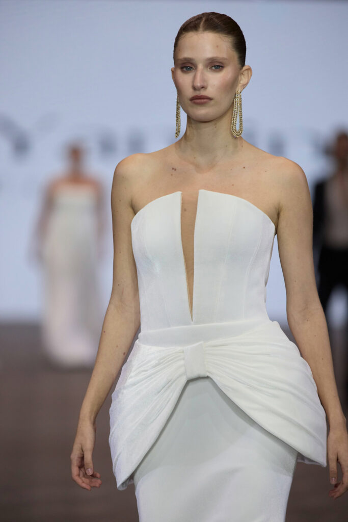 דוגמנית צועדת על המסלול בשמלה לבנה ללא כתפיות עם מחשוף עמוק ופרט קשת גדול במותניים, עונדת עגילים ארוכים מתנדנדים. הרקע מטושטש עם דוגמניות אחרות ורקע בהיר. - Liel & Cavalli