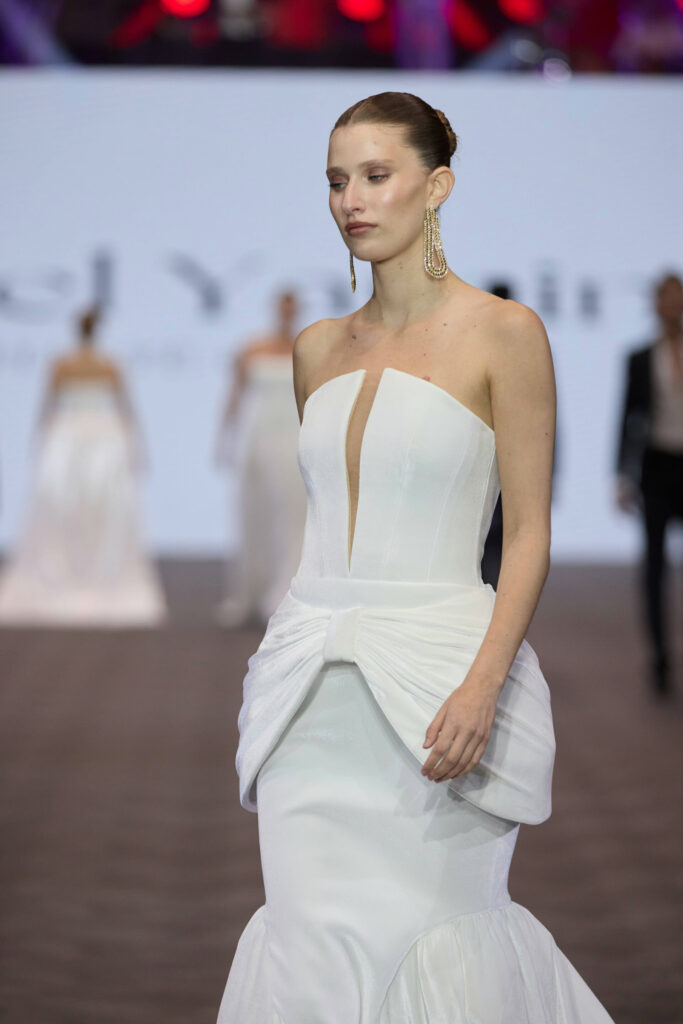 דוגמנית צועדת על המסלול בשמלה לבנה ללא כתפיות עם חתך קדמי ופרט קשת גדול במותניים. היא עונדת עגילים ארוכים ומתנדנדים ושיערה אסוף לאחור. הרקע מטושטש וניתן לראות דוגמניות אחרות. - Liel & Cavalli