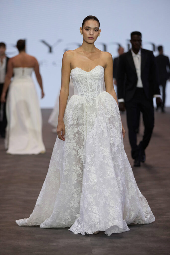 דוגמנית צועדת על מסלול תצוגה לבושה בשמלת כלה לבנה ללא כתפיות, עם מחוך צמוד וחצאית מלאה. ברקע נראות דוגמניות אחרות לבושות בבגדים רשמיים. - Liel & Cavalli