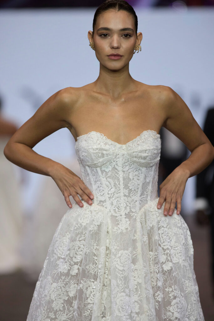 דוגמנית מדגמנת על מסלול תצוגה לבושה בשמלת כלה לבנה ללא כתפיות, עם מחוך צמוד וחצאית מלאה. שיערה אסוף לאחור, וידיה מונחות על מותניה על רקע מטושטש. - Liel & Cavalli