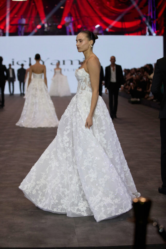 דוגמנית צועדת על מסלול תצוגה לבושה בשמלת נשף לבנה ללא כתפיות עם חצאית מלאה. ברקע נראים דוגמניות אחרות בשמלות דומות וקהל. הסצנה מתרחשת תחת אורות בוהקים ווילונות אדומים. - Liel & Cavalli