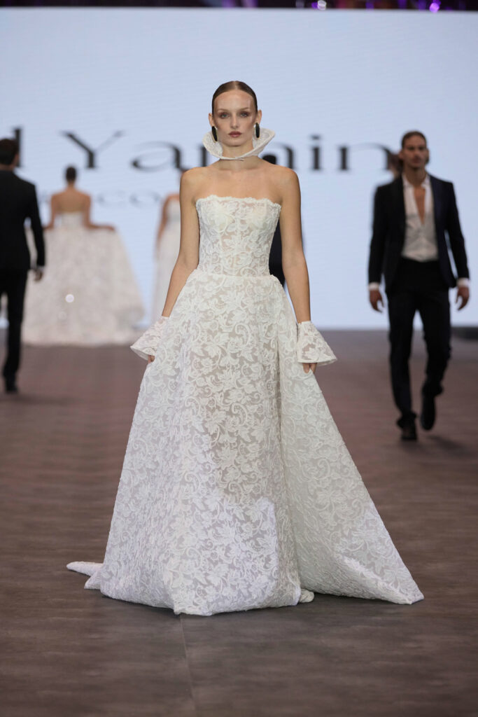 דוגמנית צועדת על מסלול תצוגה לבושה בשמלת נשף אלגנטית, ללא כתפיות, עם חצאית ארוכה ושרוולים מתרחבים ייחודיים, על רקע דמויות מטושטשות ומסך גדול. - Liel & Cavalli