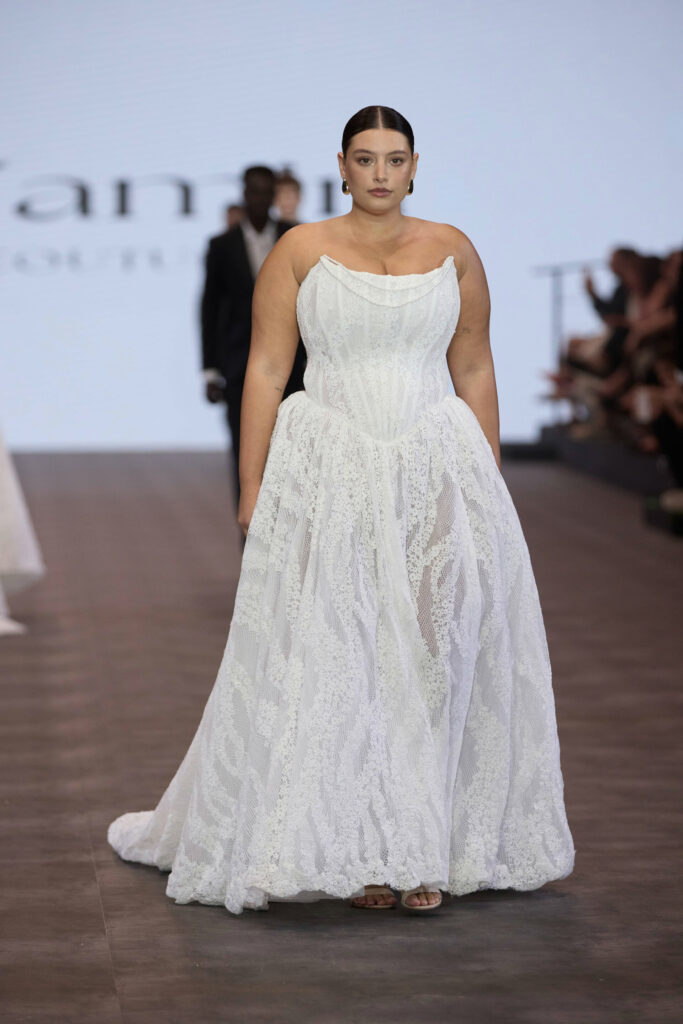 דוגמנית לבושה בשמלת כלה לבנה, ללא כתפיות, עם תחרה, צועדת על מסלול תצוגה. שיערה אסוף לאחור, וברקע נראים קהל מטושטש ודוגמנית נוספת לבושה בחליפה. - Liel & Cavalli