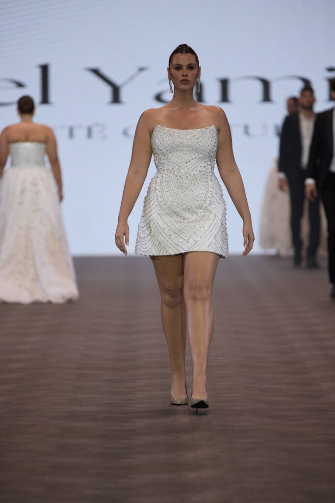 דוגמנית צועדת על מסלול תצוגה לבושה בשמלה קצרה, ללא כתפיות, עם חרוזים לבנים. ברקע נראים דוגמניות אחרות ומסך גדול עם טקסט חלקי. - Liel & Cavalli