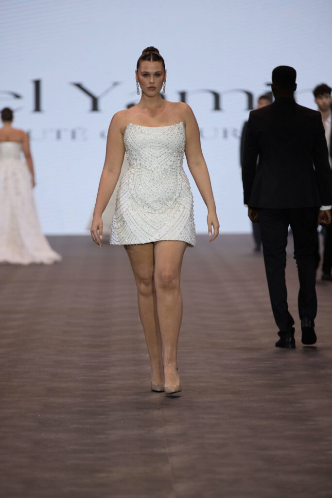 דוגמנית צועדת על מסלול תצוגה לבושה בשמלה קצרה, ללא כתפיות, עם חרוזים לבנים ועקבים שקופים. דוגמניות אחרות נראות על המסלול, וברקע מוצג חלק משמו של מעצב על מסך. - Liel & Cavalli