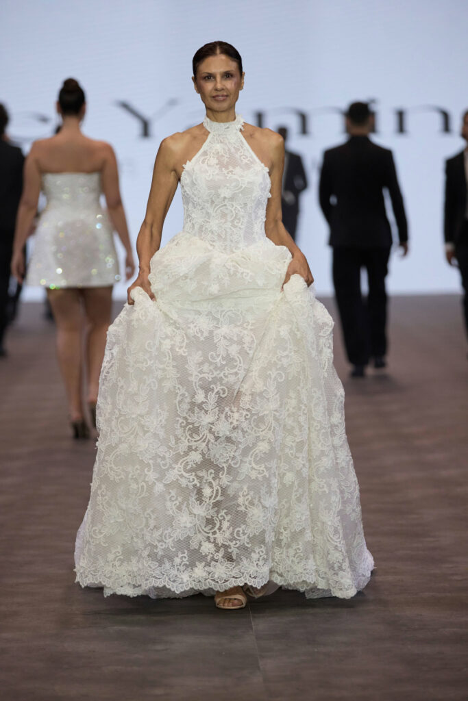 דוגמנית צועדת על מסלול תצוגה לבושה בשמלת כלה לבנה, מתחרה, ללא שרוולים, עם מחשוף גבוה, אוחזת בשולי החצאית, בעוד דוגמניות אחרות נראות ברקע. - Liel & Cavalli