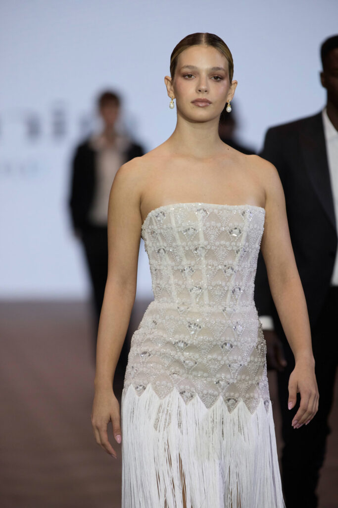 דוגמנית צועדת על המסלול בשמלה לבנה ללא כתפיות, מעוטרת בחרוזים ובפרנזים, עונדת עגילי טיפה, עם הבעה ניטרלית. ברקע נראים שני אנשים בחליפות כהות, מטושטשים. - Liel & Cavalli