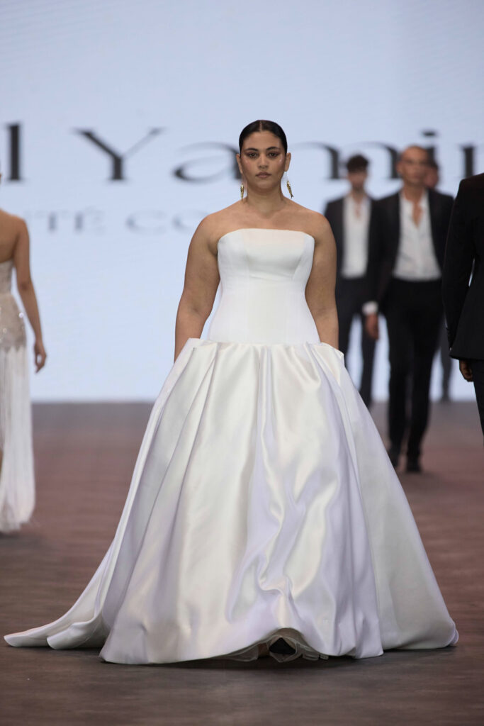 דוגמנית בשמלת כלה לבנה ללא כתפיות צועדת על מסלול תצוגה. הרקע מואר על ידי מסך גדול, ומאחוריה נראות דוגמניות אחרות בבגדי ערב. - Liel & Cavalli
