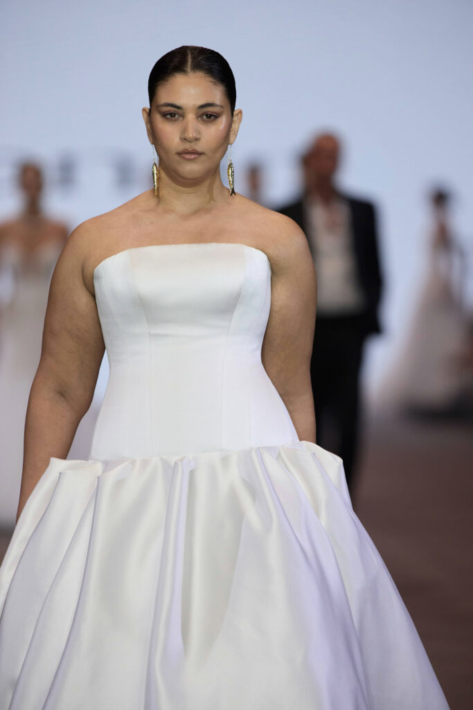 דוגמנית עם שיער משוח לאחור צועדת על המסלול בשמלת ערב לבנה ללא כתפיות, עם מחוך צמוד וחצאית מלאה, ועונדת עגילי זהב ארוכים. הרקע מטושטש וניתן לראות בו דוגמניות ואנשים אחרים. - Liel & Cavalli