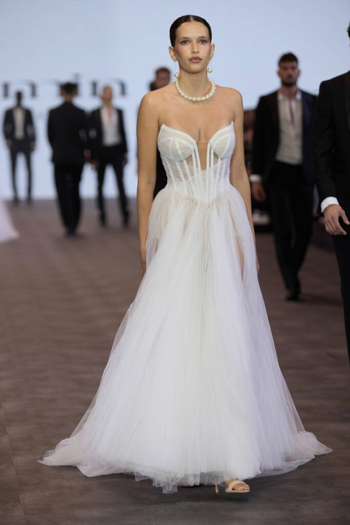 דוגמנית צועדת על מסלול תצוגה לבושה בשמלת טול לבנה ללא כתפיות, עם מחוך מבנה וצווארון לב. שיערה אסוף לאחור, היא עונדת עגילים גדולים ושרשרת פנינים; ברקע נראים אנשים מטושטשים. - Liel & Cavalli