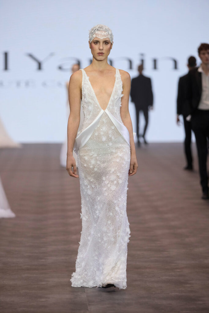 דוגמנית צועדת על מסלול תצוגה לבושה בשמלה לבנה שקופה ללא שרוולים, עם עיטורים פרחוניים וצווארון V עמוק, בשילוב עם כובע ים לבן מרקם. ברקע המטושטש נראים כמה אנשים. - Liel & Cavalli