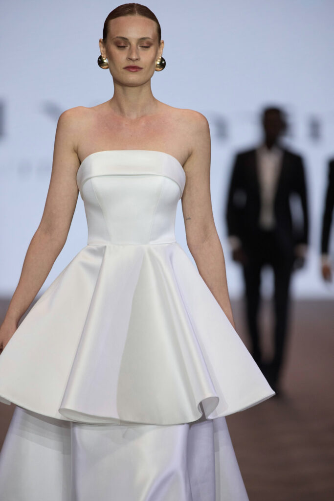 דוגמנית עם שיער משוח לאחור ועגילי זהב צועדת על המסלול בשמלה לבנה מחויטת ללא כתפיות, עם מחוך צמוד וחצאית מתנפנפת. הרקע מטושטש, וניתן לראות אנשים מרחוק. - Liel & Cavalli