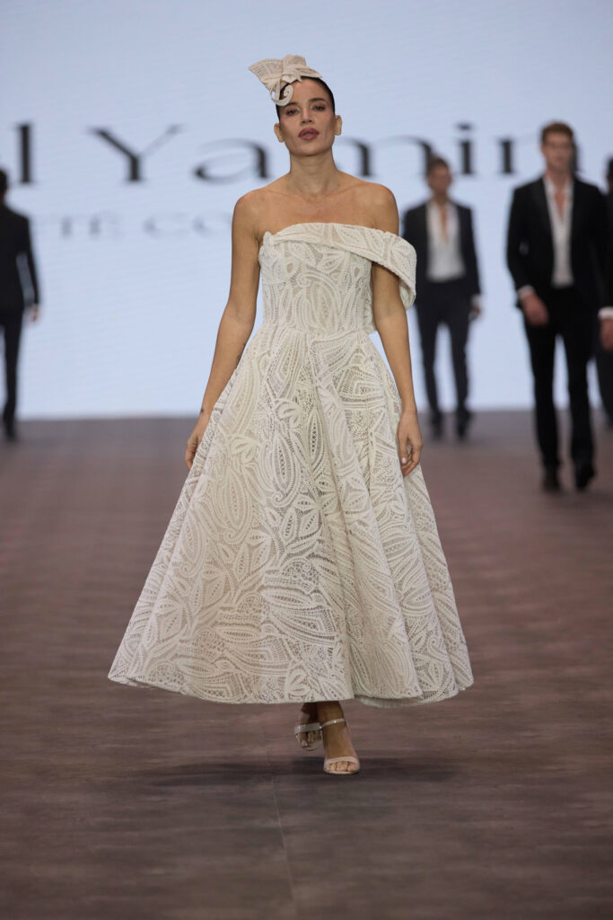 דוגמנית צועדת על המסלול בשמלת תחרה לבנה באורך הברך עם כתפיים חשופות, מעוטרת בדוגמת עלים, וכובע תואם. היא נועלת נעלי עקב בצבע גוף, וברקע המטושטש נראים כמה אנשים בחליפות. - Liel & Cavalli