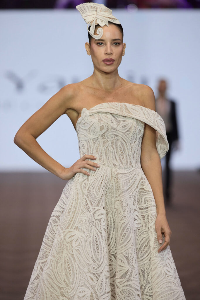 דוגמנית צועדת על המסלול בשמלת תחרה לבנה עם כתפיים חשופות, מחשוף א-סימטרי וכובע לבן מפוסל, על רקע מטושטש של תצוגת אופנה. - Liel & Cavalli