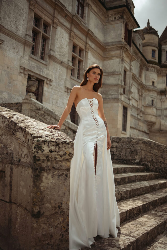 אישה בשמלה לבנה ללא כתפיות עם פרטים קדמיים משורשרים עומדת על מדרגות אבן מחוץ לבניין היסטורי, מביטה למרחק. - Liel & Cavalli