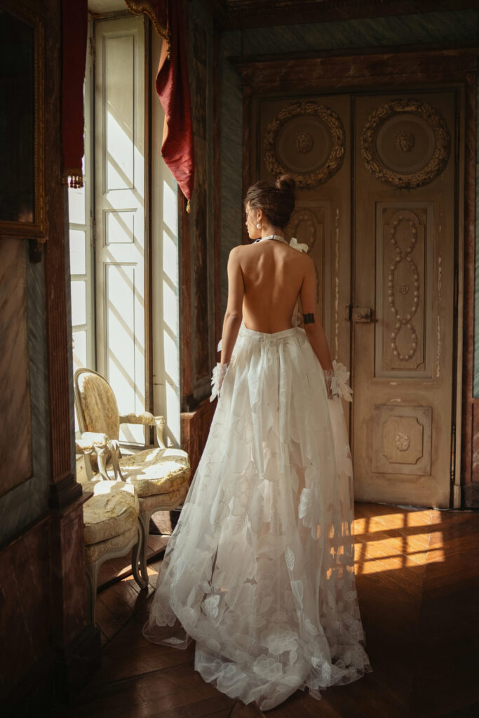 אישה בשמלה לבנה, זורמת וחסרת גב, עומדת בחדר מואר ושופע קישוטים עם רצפות עץ, ומביטה מבעד לחלון גבוה לצד כיסא אלגנטי. - Liel & Cavalli