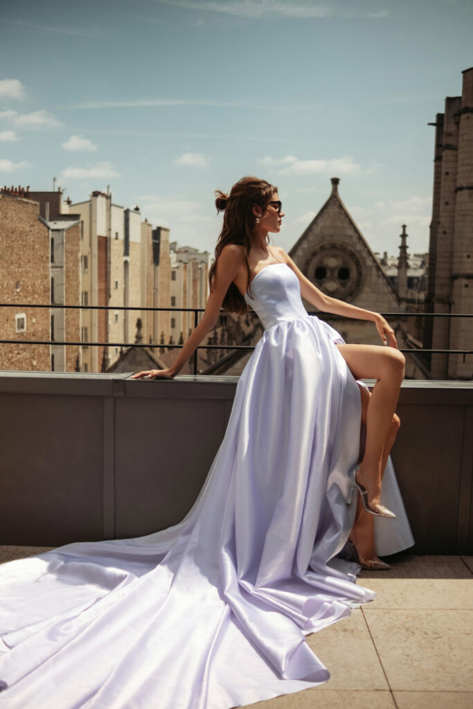 אישה בשמלה לבנה וזורמת ללא כתפיות יושבת על אדן גג, מביטה לצד. היא חובשת משקפי שמש, וברקע נראים בניינים וכנסייה בסגנון גותי תחת שמיים שטופי שמש. - Liel & Cavalli