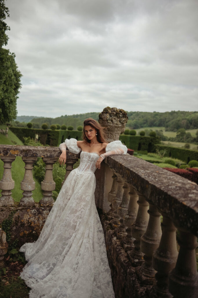אישה בשמלת כלה תחרה עם כתפיים חשופות נשענת על מעקה אבן ישן בגן ירוק ושופע, עם גבעות מתגלגלות ושמיים מעוננים ברקע. - Liel & Cavalli