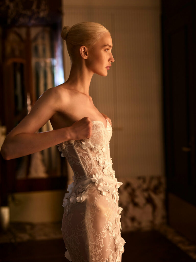 אישה עם שיער בלונדיני אסוף לקוקו מסדרת שמלה ללא כתפיות, שקופה, מעוטרת בפרטי פרחים תלת-ממדיים, בעודה עומדת בתוך הבית בתאורה חמימה ורכה. - Liel & Cavalli