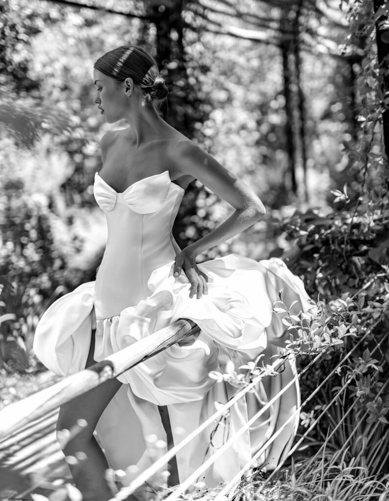 אישה בשמלה לבנה אלגנטית ללא כתפיות עומדת בחוץ בין צמחייה עבותה, מחזיקה את חצאית שמלתה ומביטה לצד. הסצנה מצולמת בשחור-לבן. - Liel & Cavalli