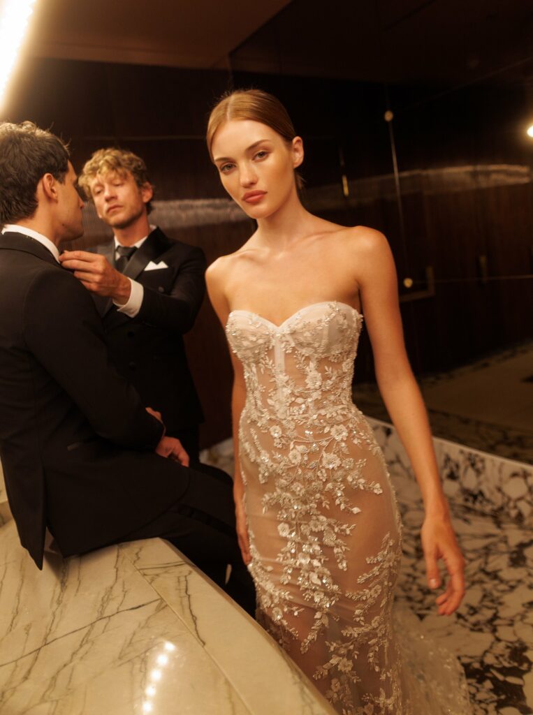 אישה בשמלה ללא כתפיות, שקופה, עם רקמת פרחים, עומדת ליד דלפק שיש. ברקע נראים שני גברים בחליפות טוקסידו, האחד מסדר את הצווארון של האחר. התפאורה כוללת קירות כהים ותאורה חמימה. - Liel & Cavalli