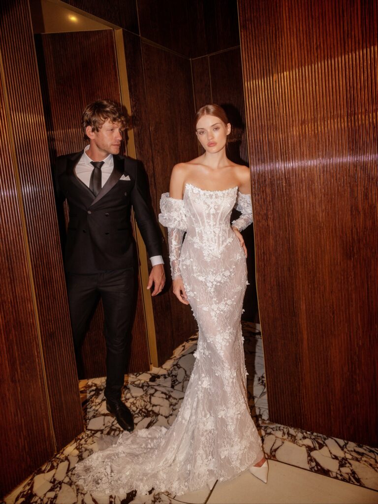 אישה בשמלת כלה אלגנטית עם כתפיים חשופות עומדת לצד גבר בחליפה שחורה ועניבה. הם נמצאים בסביבה יוקרתית עם לוחות עץ כהים ורצפות שיש. - Liel & Cavalli