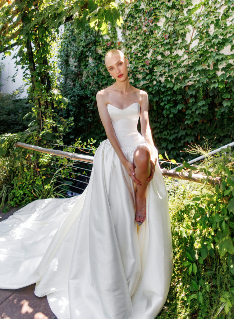 אישה בשמלת כלה לבנה ללא כתפיות יושבת בחינניות על מעקה מוקפת בעלווה ירוקה עבותה וקירות מכוסים קיסוס, כשקרני השמש מסננות מבעד לעלים. - Liel & Cavalli