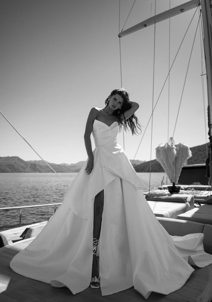 אישה בשמלה לבנה ללא כתפיות, מתנפנפת ברוח, עומדת בביטחון על סיפון יאכטה, עם הרים ומים ברקע תחת שמיים בהירים. התמונה בשחור-לבן. - Liel & Cavalli