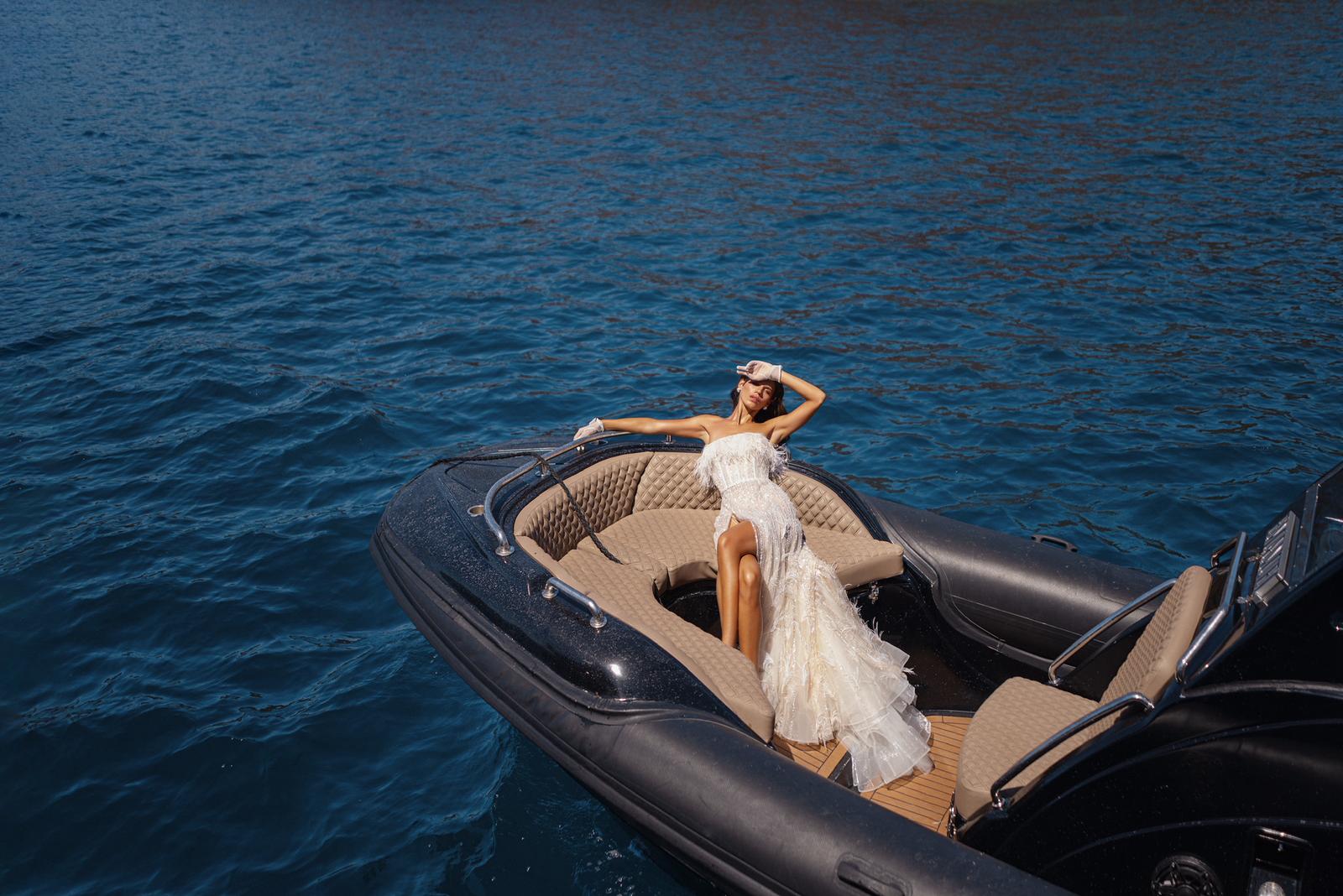 אישה בשמלה לבנה נחה על חרטום סירה שחורה הצפה על מים כחולים וצלולים, מתענגת על אור השמש, זרוע אחת מונחת על דופן הסירה והשנייה מסוככת על עיניה. - Liel & Cavalli