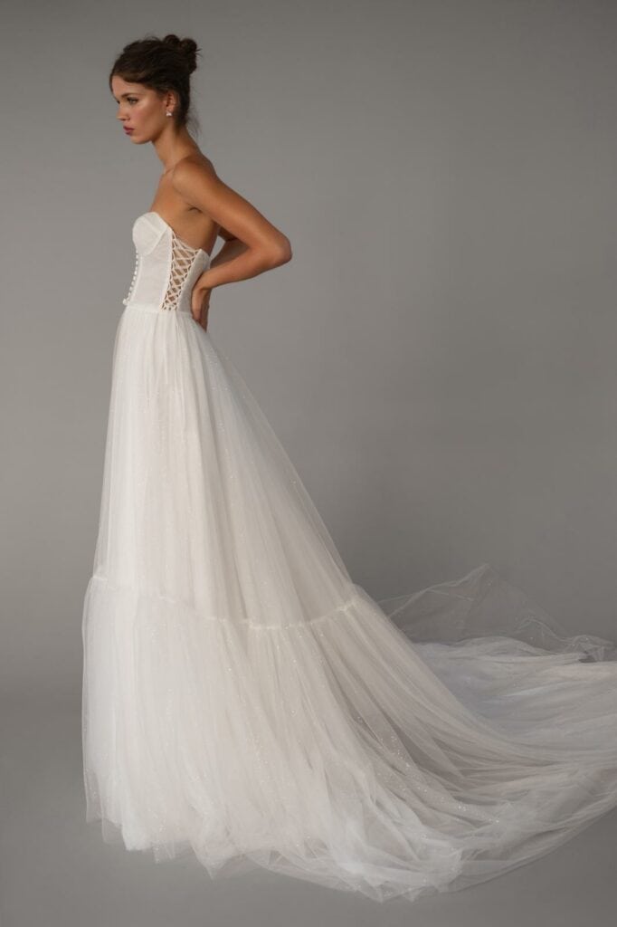 אישה לבושה בשמלת כלה לבנה ואלגנטית ללא כתפיות, עם מחוך צמוד, שרוולים עם שרוכים בצדדים ושובל טול ארוך וזורם, עומדת בפרופיל על רקע אפור אחיד. - Liel & Cavalli