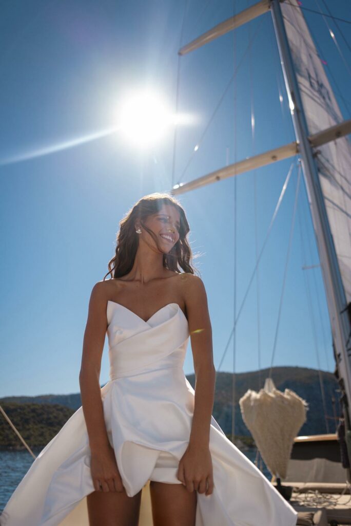 אישה בשמלה לבנה ללא כתפיות עומדת על סירה באור שמש בהיר, מחייכת עם שיער פזור. המפרש והתורן נראים מאחוריה, עם שמיים כחולים וגבעות מרוחקות ברקע. - Liel & Cavalli